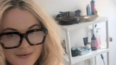 Mimmy Stranger Mimmy Stranger, with long blonde hair, and glasses, standing by a shelf, with a pigeon on top of a make-up bag, on a white shelf. She is looking straight at the camera. 