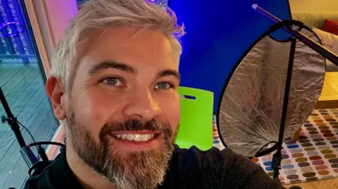 Paul Woodward Paul Woodward with grey hair and beard takes a selfie in a room with filming equipment behind him. A table and sofa is in the background with patio doors leading to decking in a garden to the left. Paul is smiling.