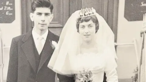 An old image of John and his wife on their wedding day. John, on the left, wears a black and white suit and has dark black hair. His wife, on the right, wears a white dress and a large vale with a silver tiara. 