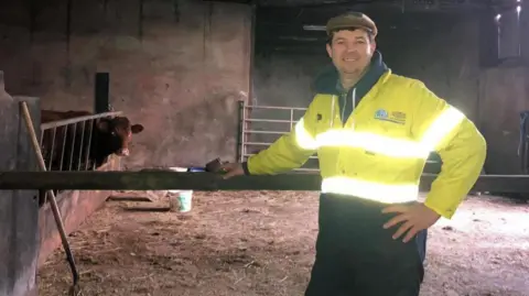 A man in a high viz yellow jacket stands in a barn. He is wearing a flat cap and is smiling a cow is behind a grill to his left. A mix of dirt and straw is on the floor