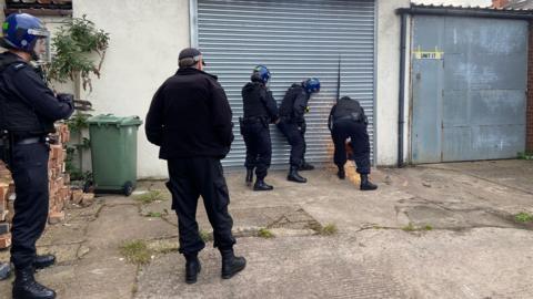 A group of police officers use a chainsaw on metal shutters of a business unit. Sparks are flying from the shutters.