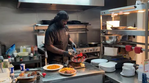 Michelle Lyons/BBC Chef Shabba is serving food from a pan onto a plate. He is wearing black overalls and gloves and has black dreadlocks. In front of him on a steel table are three plates with Caribbean food on.