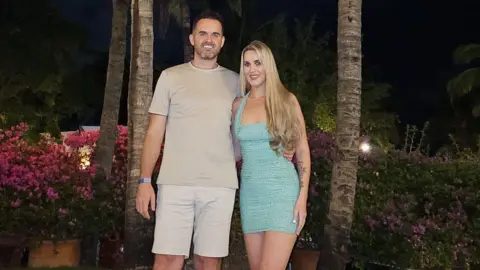 Family handout Liam is wearing a beige t-shirt and beige shorts and Natasha is wearing a light blue dress. They are standing in front of palm trees at night time.