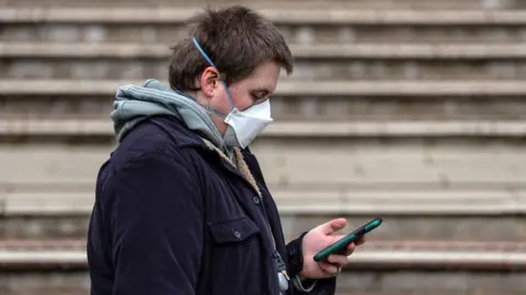 A young person wearing a hoodie, jacket and a mask across his face reads his phone.