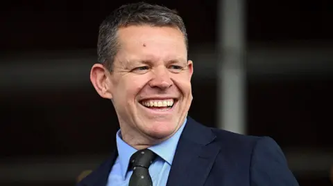 Rhun ap Iorwerth in a dark blue suit and a light blue shirt, wearing a dark green and pink dotted tie. It is a head and shoulders shot.