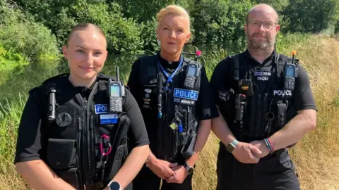 Sam Read/BBC Three volunteer police officers, standing in a row, with their arms folded in front of them. They are all wearing black trousers, and a black police jacket. Pete, on the right, has a bald head, glasses on and a short beard. Lorna, in the middle, has long blonde hair tied back and Sophie, left, has her hair in a centre parting, tied back. They are all looking at the camera. They are by water with trees behind them and grasses close to them. 