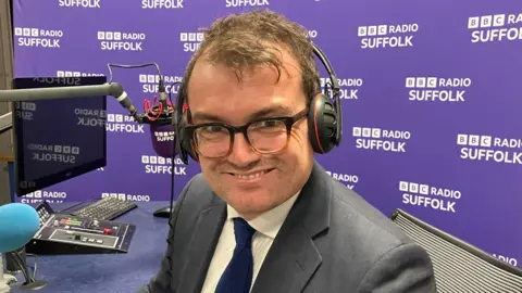 Vikki Irwin/BBC A man sits in a radio studio and smiles at the camera. He wears headphones, a suit, shirt, tie and glasses.