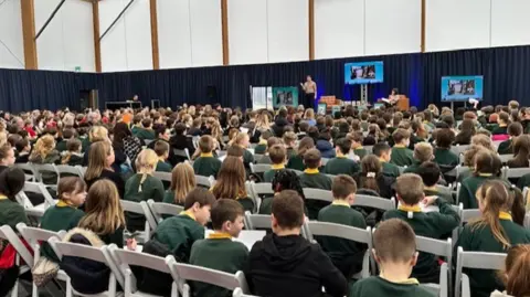A large gathering of children in chairs looking at a stage in the distance with a man with his hand in the air