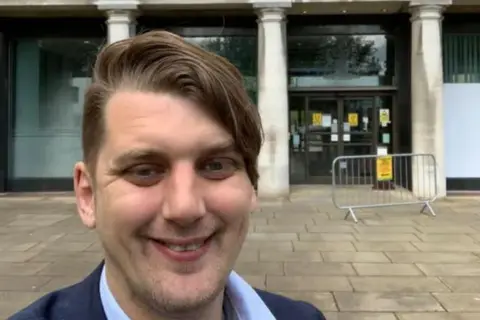 Johno Lee Image of a man with brown hair in front of a council building. He is smiling for the camera.