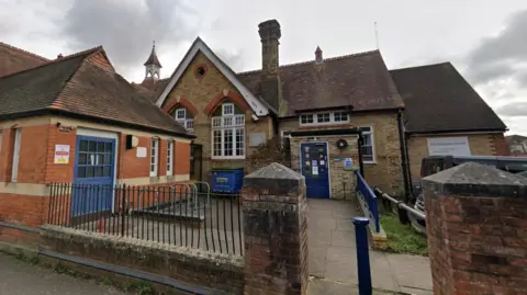 Google The Maybury Centre Trust building, made up of red and brown bricks with blue blues, stands below a grey and cloudy sky. A brick fence with black metal bars sits in front of the building.