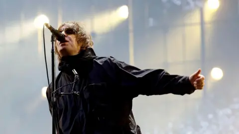 BBC Liam Gallagher wears a black raincoat and sunglasses as he tilts his head upwards to sing into a microphone on stage. Spotlights are shining behind him. He is holding his arm out with his thumb sticking up as he sings.