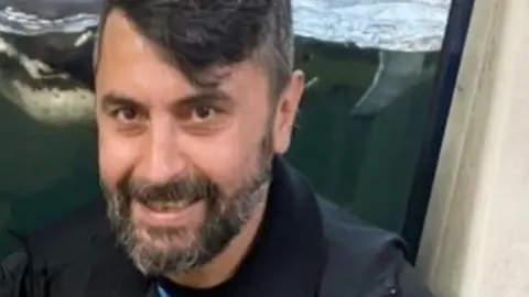 Essex Police Cumali Turhan has black hair swept to one side and a greying, black beard. He is wearing a black jacket and appears to be sitting in front of a penguin enclosure at a zoo.
