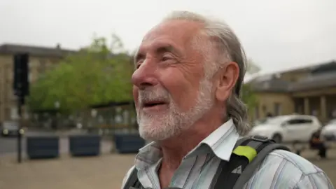 Robert stands in front of the camera. He is an elderly man with silver stubble on his chin and slicked-back hair.