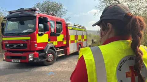 BBC A person standing with their back to the camera wearing a red top, yellow hi-vis with the British Red Cross badge on the back and a grey cap. They have long red hair which is plaited. Next to them is a parked red and yellow fire engine.