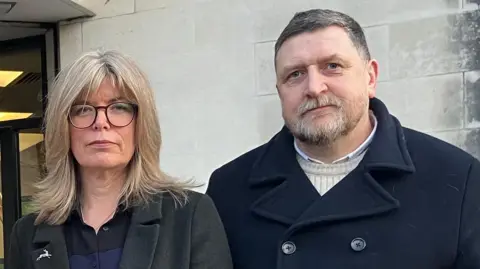 Liz Hopkin (left) on the right with blonde hair and glasses, standing outside Swansea Crown Court with her husband John, on the right. He has got short grey hair, a beard, and is wearing a dark navy coat.