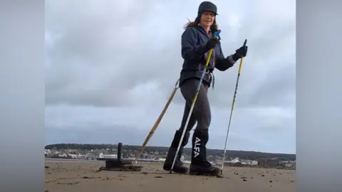 A woman with long brown hair and hiking boots is dragging a sled along a beach with the Grand Pier in Weston-super-Mare in the background. The sled is connected to her waist. She is holding walking poles.