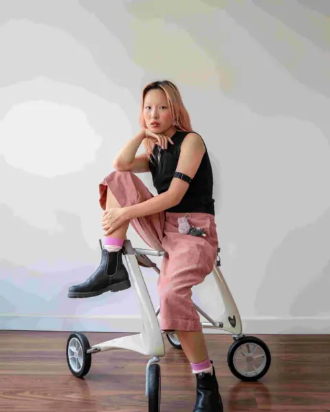 Jo Tang The image shows Josie Tang with pink hair. She is sitting on a wheeled mobility walker in a studio, wearing a black sleeveless top, pink trousers and black boots.
