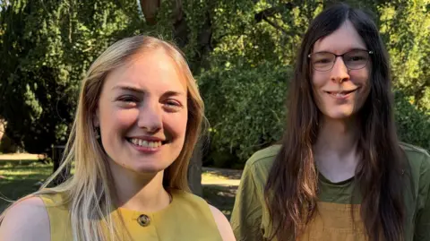 Two women standing in a garden with mature trees, lawns and buildings visible in the background in St Albans. Both are smiling.