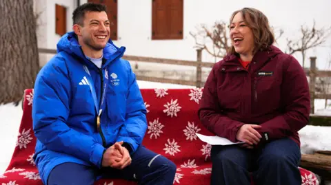 PA A woman with brown shoulder length hair wearing a burgundy coat is sat on a bench covered in a red blanket, next to a man wearing a bright blue 'Team GB' coat with a gold Olympic medal around his neck. They are both smiling.