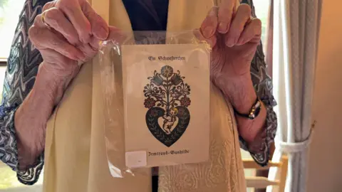 An elderly lady holding on to a piece of paper in a plastic wrapper with a decorative design and German text