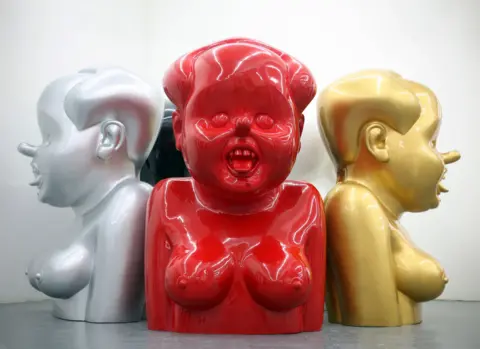 BBC/Supplied: Gao Qiang Three identical sculptures, one silver, one red, and one gold, resemble a caricature of Mao Zedong with large exposed breasts, an open mouth, a long nose and wide eyes
