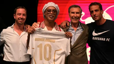 Ronaldinho holds a shirt bearing his name at a presentation event, he has one man to his right and two to his left