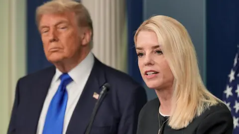 US Attorney General Pam Bondi, a woman with short blonde hair wearing a black blouse, speaks in front of a US flag, with US President Donald Trump behind  her wearing a dark blue jacket, white shirt and bright blue tie.