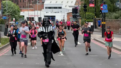 Great Birmingham Run A person dressed as Darth Vader from Star Wars is running in front of other men and women, who are also competing but wearing shorts and T-shirts.