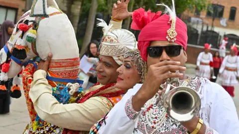 Pacemaker A man is playing a silver trumpet. He is wearing a white top with a red turban. Another man holding a plastic horse is on the left side of the image with a woman in the middle. 