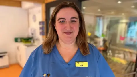 A woman in blue medical scrubs smiles at the camera. She has straight brown hair and a yellow name badge that says "Jo". There is a kitchen behind her.