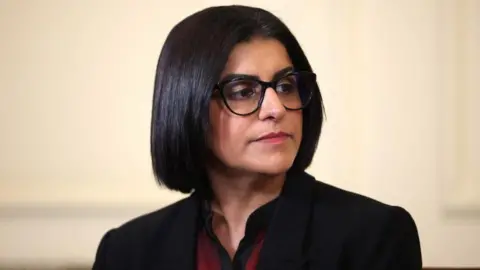 Home Secretary Shabana Mahmood is pictured looking off camera. She is wearing a dark jacket with a red and black top on beneath. 