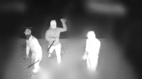 Avon and Somerset Police Black and white CCTV still showing three figures, with the rest of the view blurred. The three people, who appear to be wearing face coverings, hats and hoods to obscure their identity, can be seen brandishing machetes as one of them throws a brick.