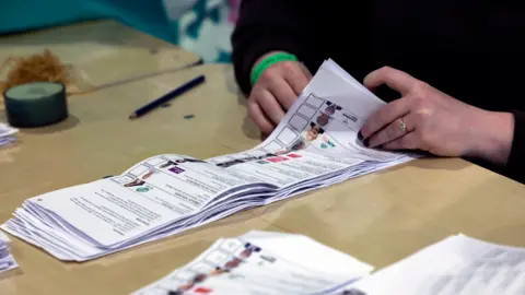 PA Media Two hands are visible flicking through a pile of ballot papers on a wooden table. 