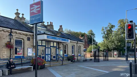 Clare Worden/BBC View of King's Lynn train station from the outside. It is a light single storey stone building with a dark grey roof. There is a blue sign which reads "King's Lynn". There are two large planters full of flowers either side of the entrance steps. 