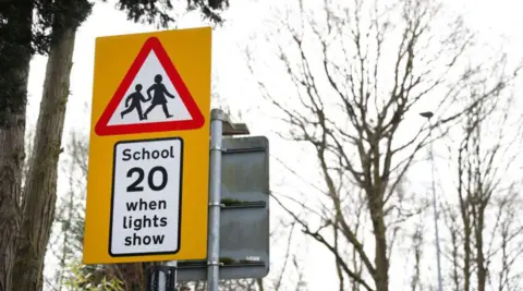 Getty Images A school road sign, including a depiction of two people within a red triangle and the words School 20 when lights show. Trees are in the background.