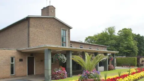 The crematorium building is on the left and in the centre of the photo. Flowers are in the foreground in front of the building and trees are on the right of the image in the background.