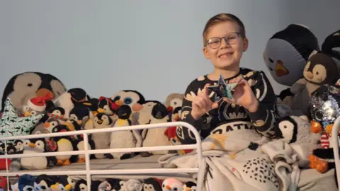 Cara Read Saxon sat on his bed surrounded by dozens of cuddly toy penguins. He is wearing batman pyjamas and is smiling while holding up a star trophy