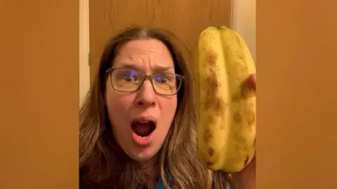 Kate Wright Kate Wright wearing glasses with brown hair looking shocked holding the "double" banana. It looks like the skin has not separated from two bananas.