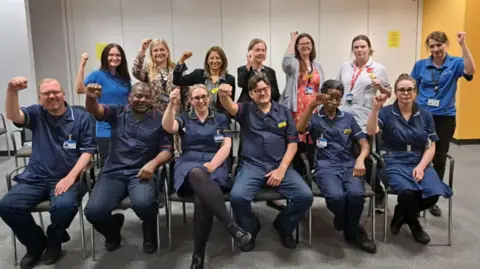 Royal Wolverhampton NHS Trust Two rows of people, some in blue uniforms, each raising an arm in the air 