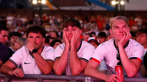 PA Media Three England fans wearing England shirts appear dejected during a screening of the World Cup 2026 semi-final match between England and Argentina at Castlefield Bowl in Manchester.