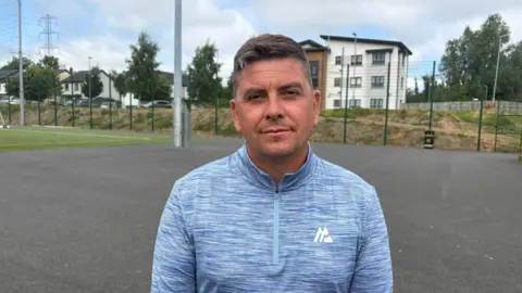 Danny Baker, with short dark hair, looks at the camera. He is wearing a light blue top. He is standing outside. Behind him is a large green fence, and behind that are some buildings. In front of the fence is a patch of grass with white sport-style markings on it. 