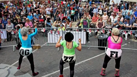 Fizzog Productions Three women are standing in front of a crowd on a high street, wearing neon coloured unitards and blonde curly wigs. They have pointed their hands in the air and are dancing. 