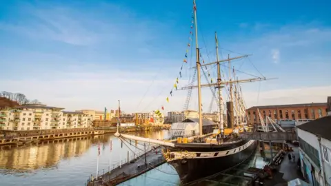 Adam Gasson/SS Great Britain The SS Great Britain, a giant black ship in Bristol Harbour, as seen from the bow. Blocks of flats and industrial buildings in a dockyard are seen around it.