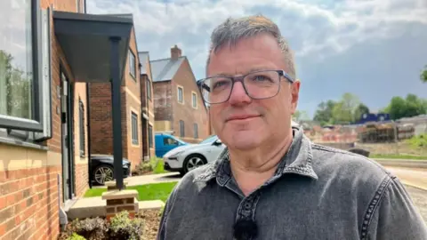 Glen Griffin, wearing glasses and a grey shirt, is standing in front of his new-build home on the adjacent estate.