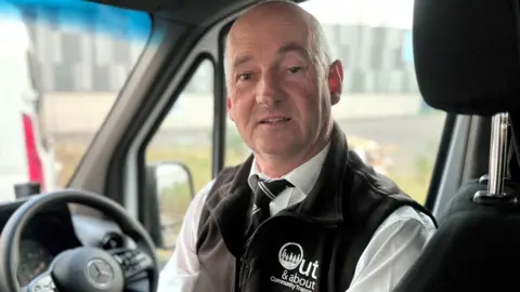 Frances Scullion sits in the driver's seat of a bus. He is turned in his seat to face the camera, which is behind him in the vehicle. In the background you can see the vehicle's steering wheel. He is wearing a black gilet, white shirt and is bald. 
