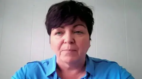 Rita Devlin, she has short dark brown hair and is wearing a blue blouse