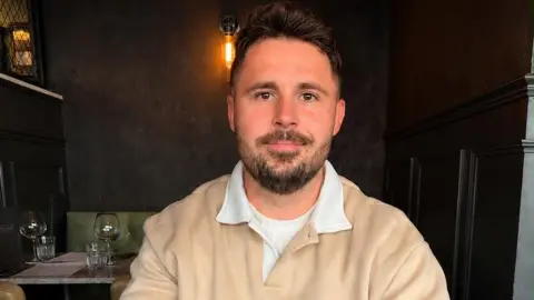 Stefan Johnson A man is looking at the camera and smiling. He is wearing a beige jumper and has a brown beard and hair. He appears to be in a restaurant.
