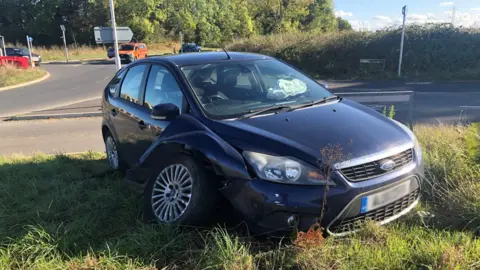 Steve Silk/BBC A dark blue Ford car has been left abandoned on a grass verge next to the roundabout, with one wheel detached from the car.