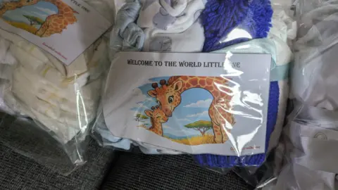 Olivia Courtney-Ashton/BBC A close-up image of see-through plastic packages, containing gifts of babygrows, baby hats and nappies. Each has a label which has a picture of a giraffe on it and the words: 'Welcome to the world little one'.