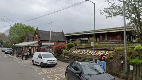The front of Westbury railway station, showing Westbury in large letters in a flower bed and a traditional red brick building, with the tracks raised up behind. A few cars in a pick-up area at the front